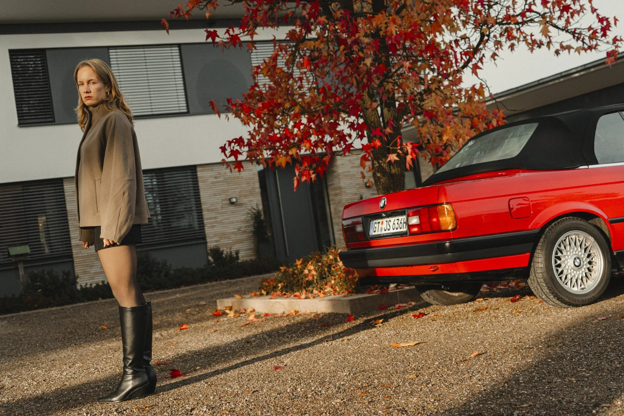 Eine Person steht neben einem roten BMW E30 vor einem Haus, daneben ein Baum mit rotem Herbstlaub.