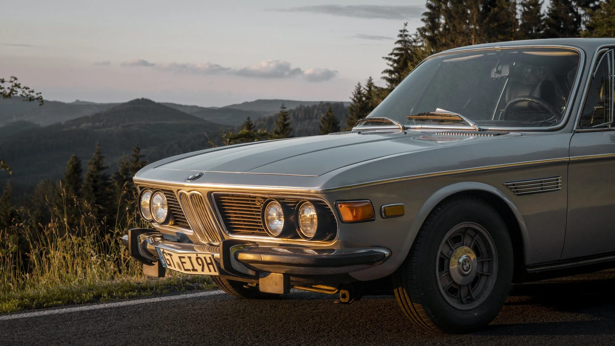 Ein silberner BMW E9 steht auf einer Straße vor einem bewaldeten Höhenzug.