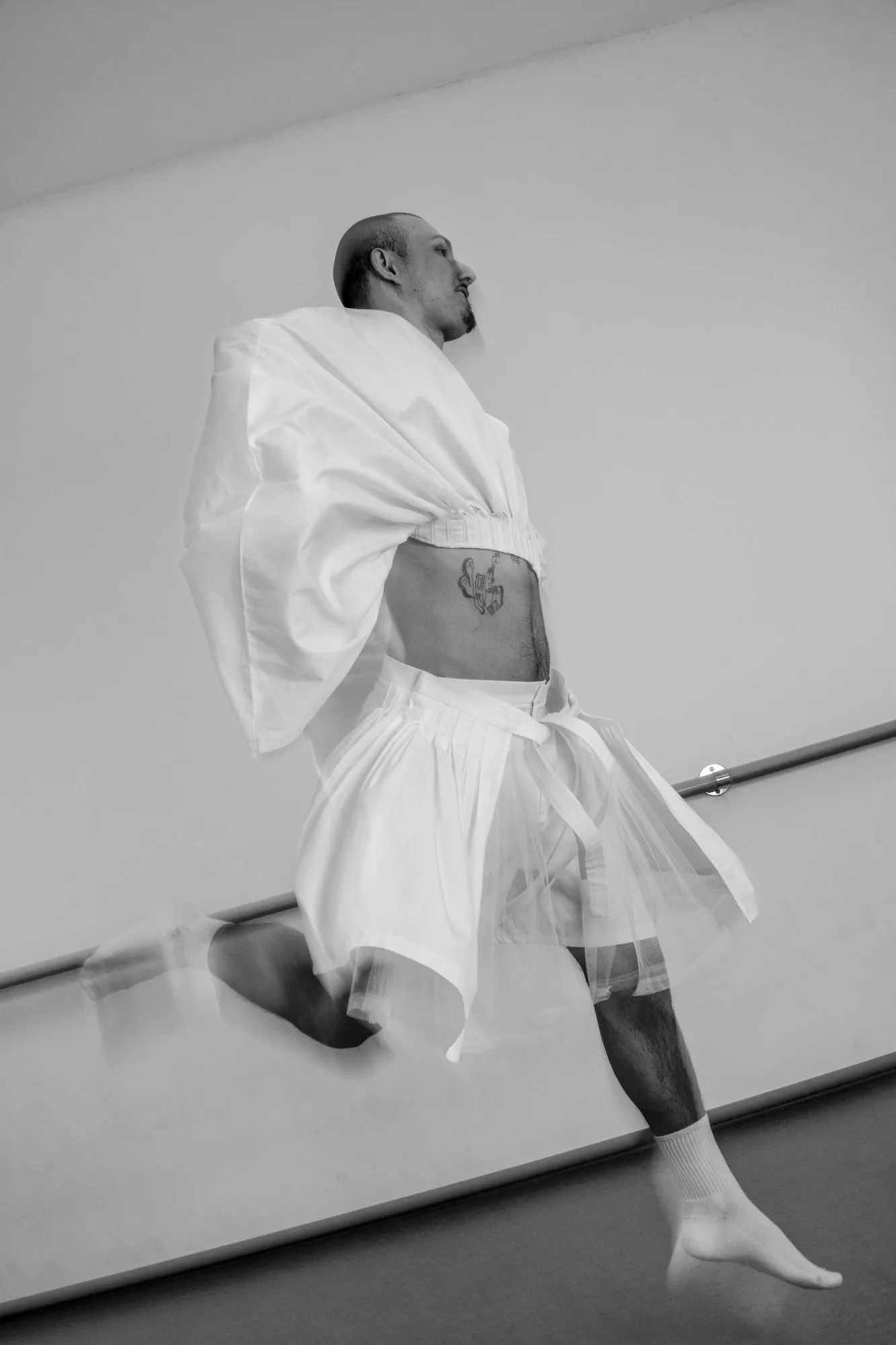 Schwarzweiß: eine Person in weißem Hemd und Tüllrock vor einer Ballettstange, ein Bein angehoben.